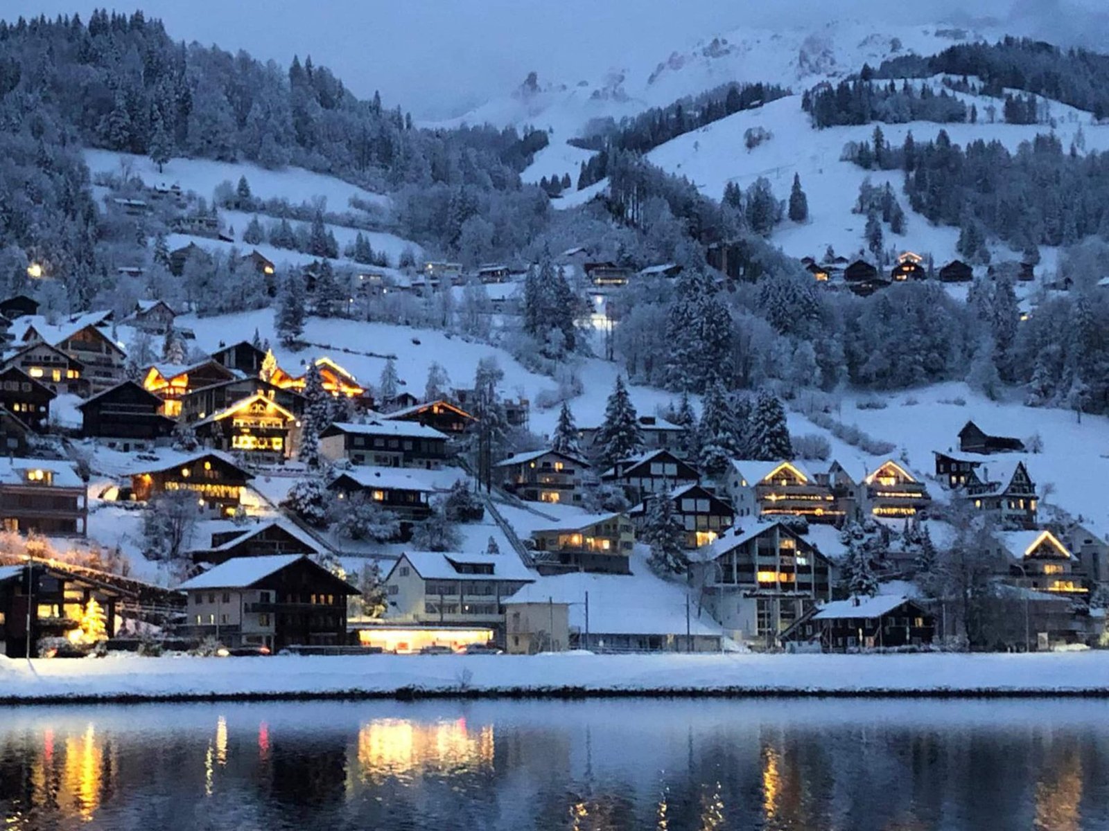 Engelberg village at dusk under snow, its lights reflected in the lake