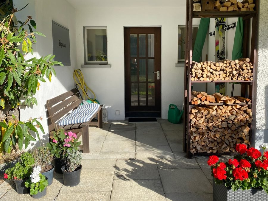 The private entrance: a dark wooden door, a bench and a stacked firewood store, geraniums in pots